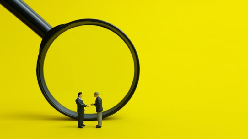 Magnifying glass over small model people shaking hands and making a deal, against bright yellow background
